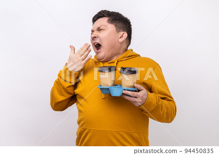 Portrait of tired exhausted man holding coffee cups and yawning, feeling bored after stressful working day, wearing urban style hoodie. Indoor studio shot isolated on white background. 94450835