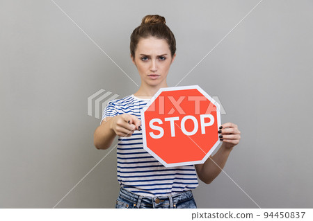 Portrait of serious bossy woman with bun hairstyle wearing striped T-shirt holding red stop sign pointing at camera, has strict expression. Indoor studio shot isolated on gray background. Portrait of serious bossy woman with bun hairstyle wearing striped T-shirt holding red stop sign pointing at camera, has strict expression. Indoor studio shot isolated on gray background. 94450837