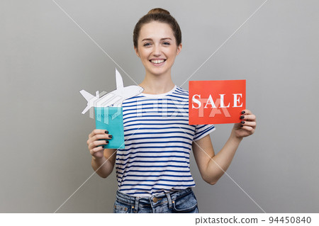 Portrait of happy woman wearing striped T-shirt holding passport with paper air plane and sale word sign, smiling at camera, traveling. Indoor studio shot isolated on gray background. 94450840