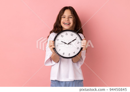 Portrait of smiling delighted little girl wearing white T-shirt holding wall clock, being happy, deadline, satisfied with completed home task. Indoor studio shot isolated on pink background. Portrait of smiling delighted little girl wearing white T-shirt holding wall clock, being happy, deadline, satisfied with completed home task. Indoor studio shot isolated on pink background. 94450845