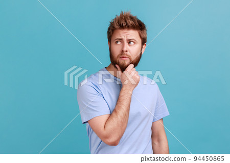 Portrait of unhappy pensive young bearded man keeping hand on chin, looking concentrated into distance, thinking about important things. Indoor studio shot isolated on blue background. 94450865