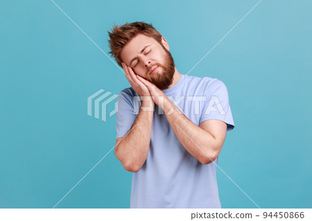Portrait of tired bearded man sleeping laying down on his palms and smiling pleased, having comfortable nap and resting, dozing off. Indoor studio shot isolated on blue background. 94450866
