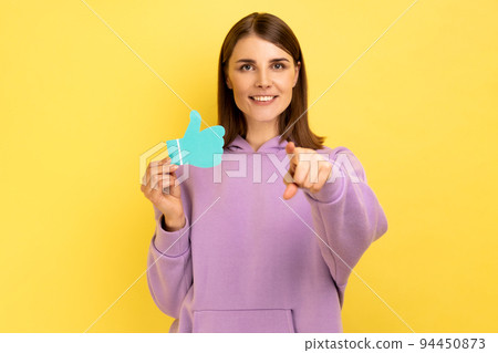 Smiling woman showing thumbs up blue paper sign, liking and recommending posts in social networks, asking to rate, pointing at camera. Indoor studio shot isolated on yellow background. 94450873