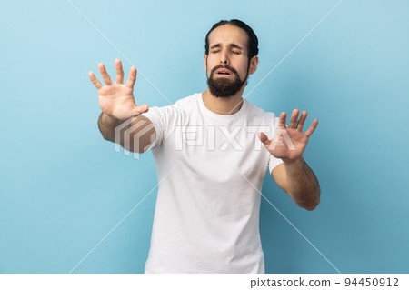 Portrait of disoriented blind man with beard wearing white T-shirt standing with stretched arms, searching for way, vision problems. Indoor studio shot isolated on blue background. 94450912