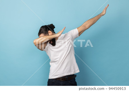 Portrait of anonymous unknown man with beard wearing white T-shirt standing in dab dance pose, internet meme, celebrating success. Indoor studio shot isolated on blue background. 94450913
