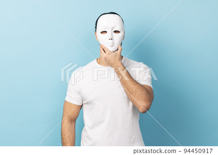 Portrait of unknown anonymous man with beard in white T-shirt covering his face with white mask, hiding personality, conspiracy and privacy, secrets. Indoor studio shot isolated on blue background. Portrait of unknown anonymous man with beard in white T-shirt covering his face with white mask, hiding personality, conspiracy and privacy, secrets. Indoor studio shot isolated on blue background. 94450917