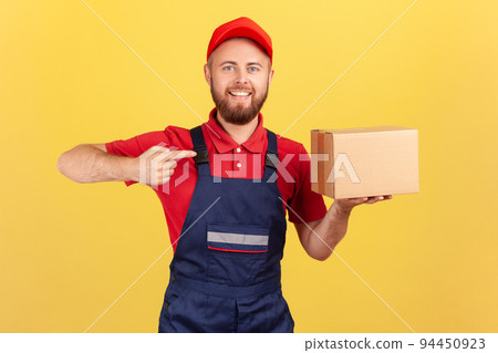 Portrait of cargo transportation, delivery service. Friendly courier man in uniform pointing at cardboard box, delivering parcel to client. Indoor studio shot isolated on yellow background. 94450923