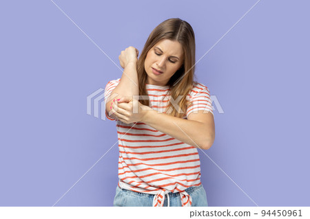 Unhealthy sick blond woman wearing striped T-shirt having trauma after car accident, feels pain in elbow, trapped nerves, rheumatoid arthritis. Indoor studio shot isolated on purple background. 94450961