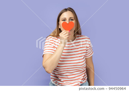 Portrait of positive blond woman wearing striped T-shirt holding little red heart and covering her mouth with love symbol. Indoor studio shot isolated on purple background. 94450964