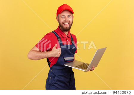 Satisfied worker man wearing blue overalls, red T-shirt and cap working on laptop, showing thumb up, likes a new online service, looking at camera. Indoor studio shot isolated on yellow background. Satisfied worker man wearing blue overalls, red T-shirt and cap working on laptop, showing thumb up, likes a new online service, looking at camera. Indoor studio shot isolated on yellow background. 94450970