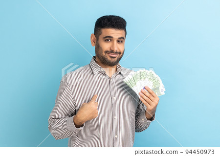 Portrait of young adult businessman with beard holding euro bills and pointing at himself with pride, looking at camera, wearing striped shirt. Indoor studio shot isolated on blue background. 94450973