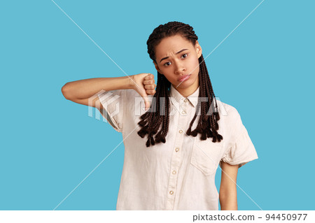 Discontent woman with black dreadlocks showing disapproval sign, keeps thumb down, expresses dislike, frowns face in discontent, wearing white shirt. Indoor studio shot isolated on blue background. 94450977