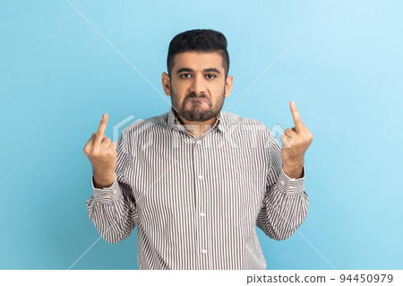 Impolite businessman showing middle fingers and asking to get off looking at camera with negativity, disrespectful behaviour, wearing striped shirt. Indoor studio shot isolated on blue background. 94450979