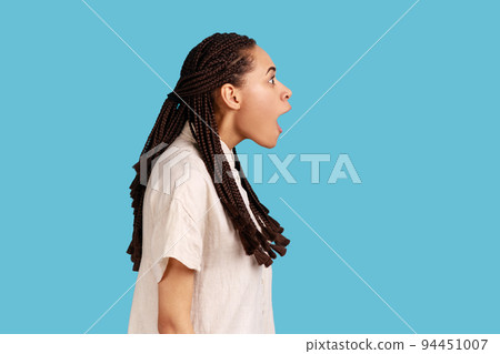 Side view portrait of happy surprised beautiful woman with black dreadlocks notices something incredible, stares amazed, keeps mouth opened. Indoor studio shot isolated on blue background. 94451007