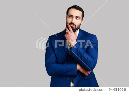 Pensive thoughtful bearded businessman holding his chin with hand, pondering about future, planning strategy, wearing official style suit. Indoor studio shot isolated on gray background. Pensive thoughtful bearded businessman holding his chin with hand, pondering about future, planning strategy, wearing official style suit. Indoor studio shot isolated on gray background. 94451016