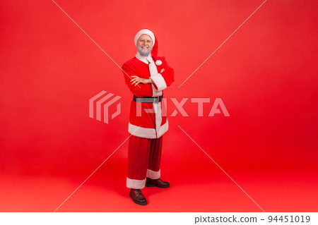 Full length photo of elderly man with gray beard wearing santa claus costume with confident look, standing with folded arms and looking at camera. Indoor studio shot isolated on red background. 94451019