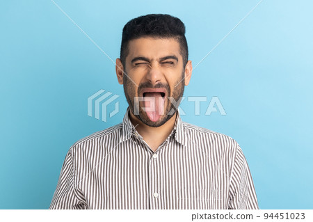Photo of young handsome foolish crazy businessman, keeping eyes closed and showing tongue out, expressing positive emotions, wearing striped shirt. Indoor studio shot isolated on blue background. 94451023
