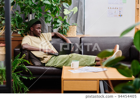 Young relaxed man in casualwear with laptop sitting on black leather couch 94454405