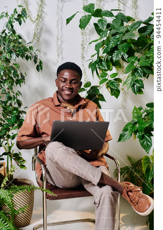 Young smiling employee in casualwear sitting on chair with laptop on his knees 94454411