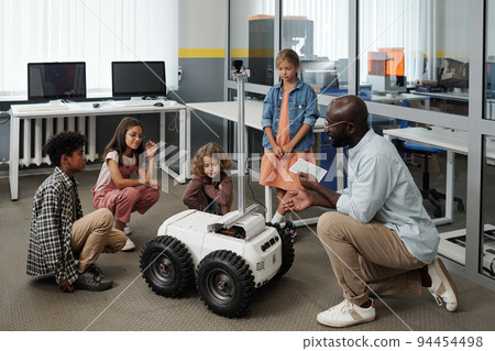Young teacher of robotics with tablet making presentation of crawler to kids Young teacher of robotics with tablet making presentation of crawler to kids 94454498