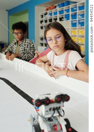 Clever youthful schoolgirl in eyeglasses looking at toy robot moving along table Clever youthful schoolgirl in eyeglasses looking at toy robot moving along table 94454521