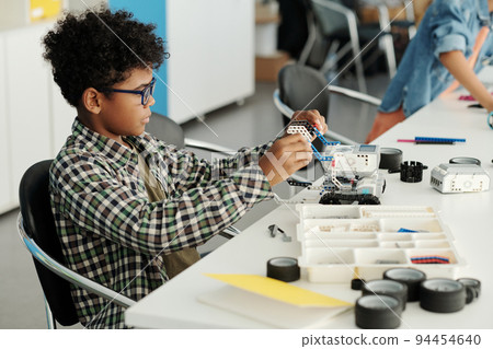 Clever African American schoolboy constructing new robot by desk at lesson Clever African American schoolboy constructing new robot by desk at lesson 94454640