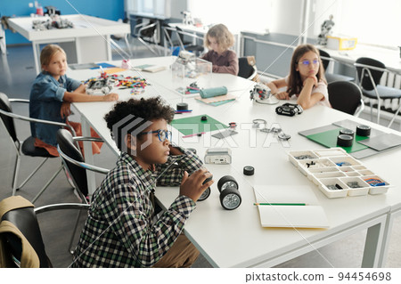 Group of intercultural schoolkids sitting by large desk during presentation Group of intercultural schoolkids sitting by large desk during presentation 94454698