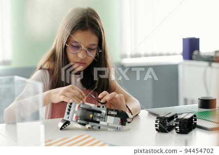 Cute serious schoolgirl creating robot or model of machine during lesson Cute serious schoolgirl creating robot or model of machine during lesson 94454702