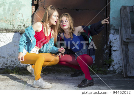 Two funny girls in bright clothes in the style of the nineties are sitting against the background of a rusty door. Russian village girls. Two funny girls in bright clothes in the style of the nineties are sitting against the background of a rusty door. Russian village girls. 94462052