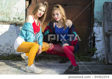 Two funny girls in bright clothes in the style of the nineties are sitting against the background of a rusty door. Russian village girls. Two funny girls in bright clothes in the style of the nineties are sitting against the background of a rusty door. Russian village girls. 94462054