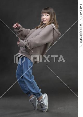Beautiful teen girl in a sweatshirt and jeans dances in front of the camera on a gray background. Beautiful teen girl in a sweatshirt and jeans dances in front of the camera on a gray background. 94464463