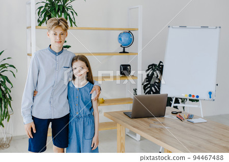 Teenage girl and boy standing near table with laptop in classroom looking to the camera. Teen schoolboy and schoolgirl do project on computer together at school Teenage girl and boy standing near table with laptop in classroom looking to the camera. Teen schoolboy and schoolgirl do project on computer together at school 94467488