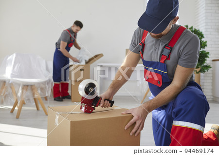 Worker from moving company or delivery service sealing cardboard box with tape dispenser 94469174