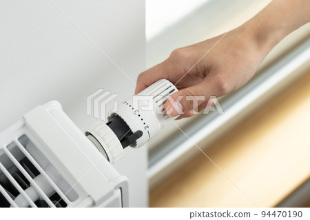 A woman adjusts the radiator thermostat to the economical mode of room heating A woman adjusts the radiator thermostat to the economical mode of room heating 94470190