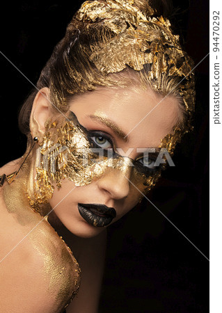 Closeup portrait of beautiful young woman with golden foil on face. Creative golden makeup on black background 94470292