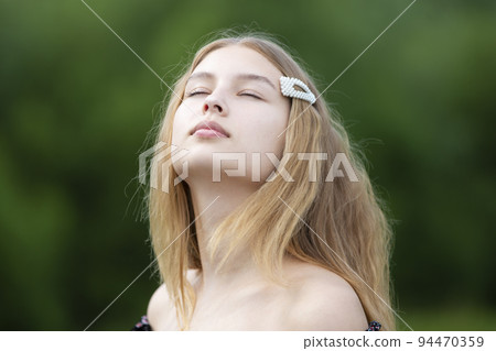 A beautiful girl with long hair closed her eyes on a summer background. The face of a fair-haired teenage girl. 94470359