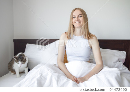 Happy young woman with cat in bed at home. Pets, morning, comfort, rest and people concept. 94472490