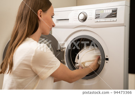 Young friendly woman with long hair, dressed in a white T-shirt, puts dirty white clothes from a basket into a white washing machine in the laundry room. 94473366