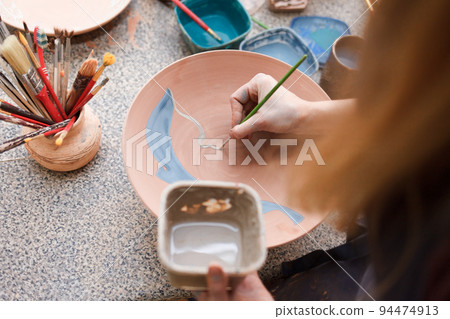 Potter woman paints a ceramic plate. Girl draws with a brush on earthenware. Process of creating clay products.. 94474913