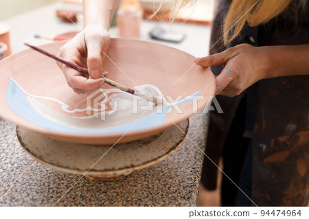Potter woman paints a ceramic plate closeup. Girl draws with a brush on earthenware. Process of creating clay products... 94474964