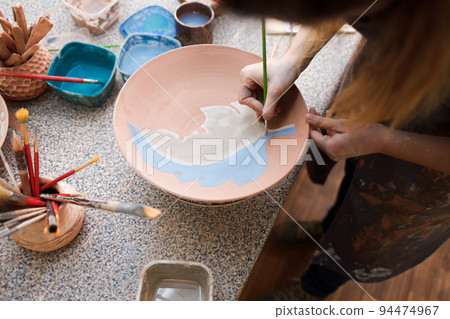 Potter woman paints a ceramic plate. Girl draws with a brush on earthenware. Process of creating clay products.. 94474967