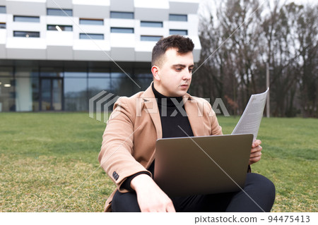 Man is studying while sitting with a notebook in his hands on the grass near the university.. Man is studying while sitting with a notebook in his hands on the grass near the university.. 94475413