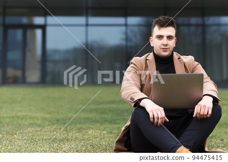 Student working on a laptop outdoor. Man sits with a laptop in his hands on the backcground of a business center.. Student working on a laptop outdoor. Man sits with a laptop in his hands on the backcground of a business center.. 94475415