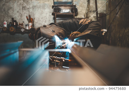 Worker in a welder mask works in a workshop for welding iron. Man makes iron products. Guy works with a welding machine using a mask to protect his eyes from dangerous rays 94475680