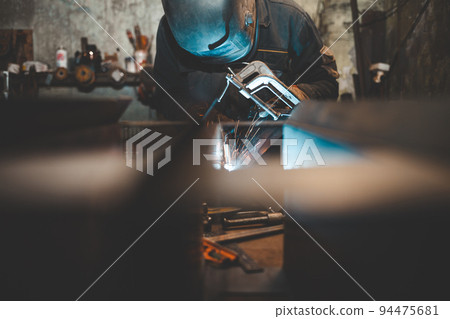Worker in a welder mask works in a workshop for welding iron. Man makes iron products. Guy works with a welding machine using a mask to protect his eyes from dangerous rays Worker in a welder mask works in a workshop for welding iron. Man makes iron products. Guy works with a welding machine using a mask to protect his eyes from dangerous rays 94475681