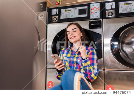 Pretty girl with smartphone in hand listens to music leaning on a washing machine in public laundry. Pretty girl with smartphone in hand listens to music leaning on a washing machine in public laundry. 94476074