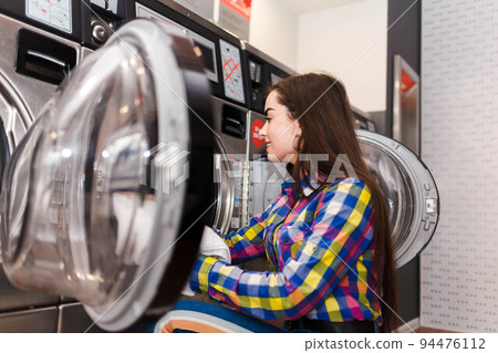 Girl loads laundry into a washing machine. woman in laundrette. Girl loads laundry into a washing machine. woman in laundrette. 94476112