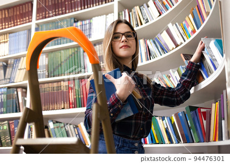 Cute student in glasses takes a book from the library bookshelf. Young lady is looking for the right literature among many books.. 94476301