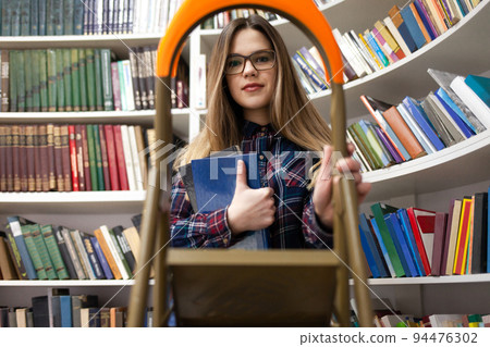 Pretty student lady stands on a stepladder in a library holding a book in her hand. Woman in public library. 94476302