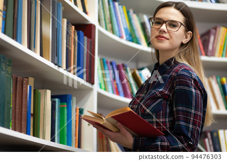 Teen with a book in hands stands in the library. Portrait of a young student in glasses which is reading a book. Teen with a book in hands stands in the library. Portrait of a young student in glasses which is reading a book. 94476303